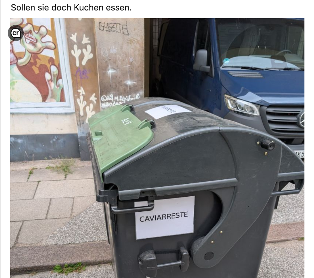 Eine Mülltonne mit grünem Deckel steht auf einem Gehweg. An der Seite der Tonne ist ein Zettel mit der Aufschrift "CAVIARRESTE" angebracht. Im Hintergrund ist ein graues Gebäude mit Graffiti und ein geparktes blaues Fahrzeug zu sehen. Oben im Bild steht der Text: "Sollen sie doch Kuchen essen." In der linken oberen Ecke ist ein kleines "CR"-Symbol.